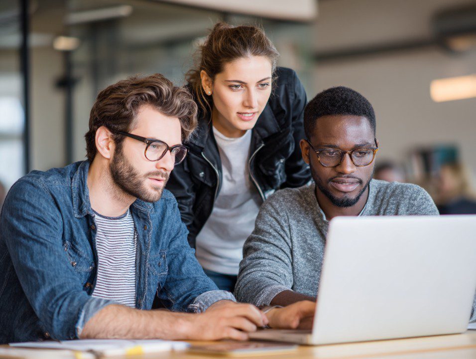 Three professionals collaborating and working together on a laptop, discussing data and problem-solving in a modern office environment