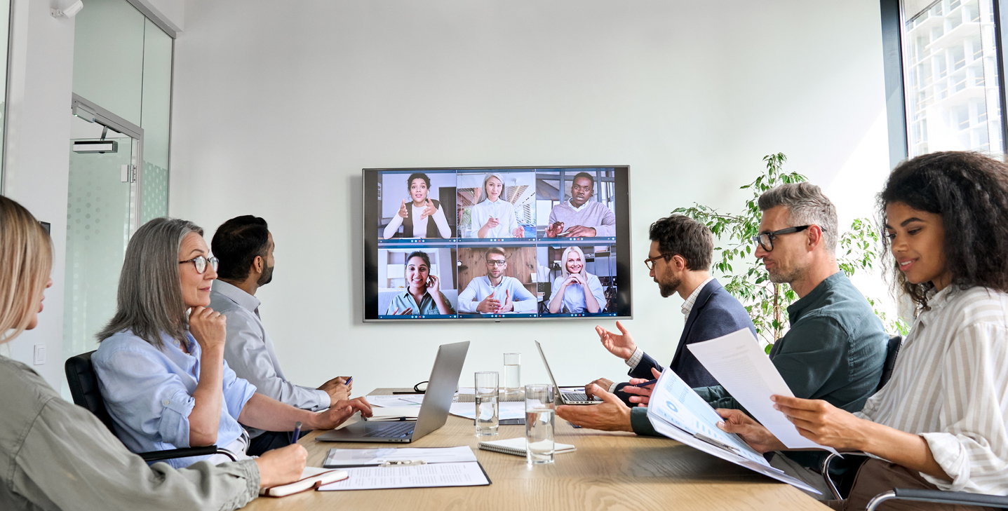 Team of professionals in a conference room attending a video call meeting on a large screen, collaborating with remote participants in a hybrid work environment