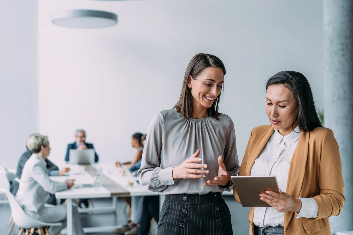 Two business professionals reviewing data on a tablet in a modern office, discussing IT solutions and digital strategy while team works in background